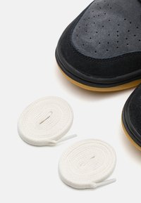 Sapatilhas de camurça pretas com design perfurado, sola de borracha preta e dois atacadores brancos enrolados com pontas de plástico.