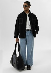 Zwarte denim oversized shirt met een witte t-shirt eronder, lichtblauwe wijdvallende jeans, zwarte lederen schoudertas en zonnebril.