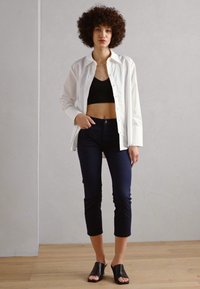 Biela košeľa na gombíky cez čierny crop top, v kombinácii s tmavomodrými priliehavými nohavicami a čiernymi šľapkami. Hladké látky a priliehavý strih.
