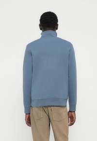 Μπλε φούτερ με ψηλό γιακά και μακριά μανίκια, κατασκευασμένο από μαλακό ύφασμα. Διαθέτει απλό σχέδιο και ριμπ τελείωμα στο κάτω μέρος και στις μανσέτες.