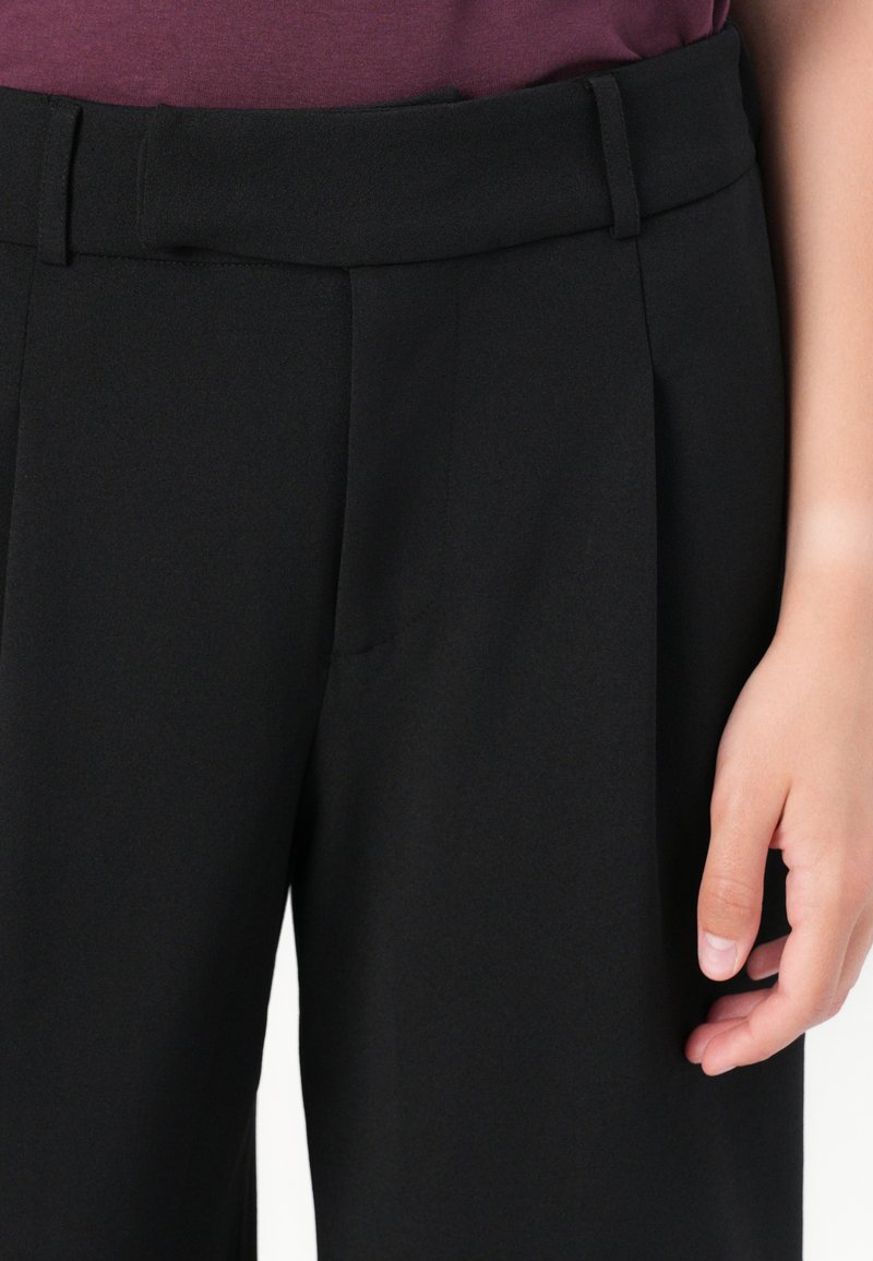 Schwarze Hose aus glattem Stoff, mit weitem Bund, vorderen Falten und Seitentaschen. Eine Hand ruht auf dem Stoff.