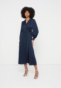 Marineblå omslagskjole med lange puffermede ermer, laget av glatt stoff, med et belte i midjen og midi-lengde, kombinert med remmesko med hæl.