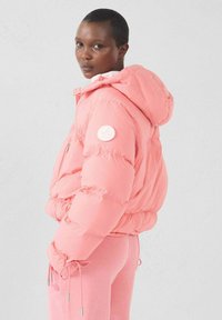 Giacca rosa imbottita con cappuccio, caratterizzata da una toppa rotonda con logo sulla manica, polsini elastici e vita aderente. Combinata con pantaloni coordinati.