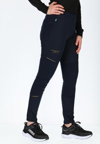 Leggings blu marina con dettagli e logo dorati, dotati di una vita alta e tasche laterali, abbinati a scarpe da ginnastica nere. Tessuto liscio ed elasticizzato.