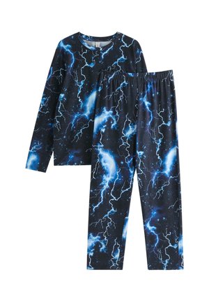 Garçon debout pieds nus portant un pyjama noir avec des motifs d’éclairs bleus et blancs et des nuages sur un fond clair uni.