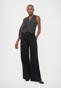 Colete cinza com riscas verticais, três botões na frente, combinado com calças largas pretas de cintura alta com bolsos tipo cargo. Sapatos pretos planos.