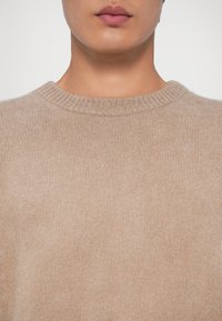 Bežinė mezginė megztinis su apvalia iškirpte, pagamintas iš minkštos medžiagos. Yra tekstūruotas paviršius ir raštuoti kraštai prie apykaklės.