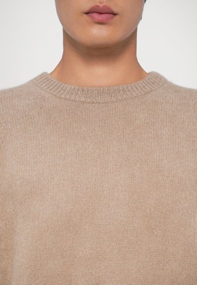 Beige pletený svetr s kulatým výstřihem, vyrobený z měkkého materiálu. Má texturovaný povrch a žebrované okraje na límci.