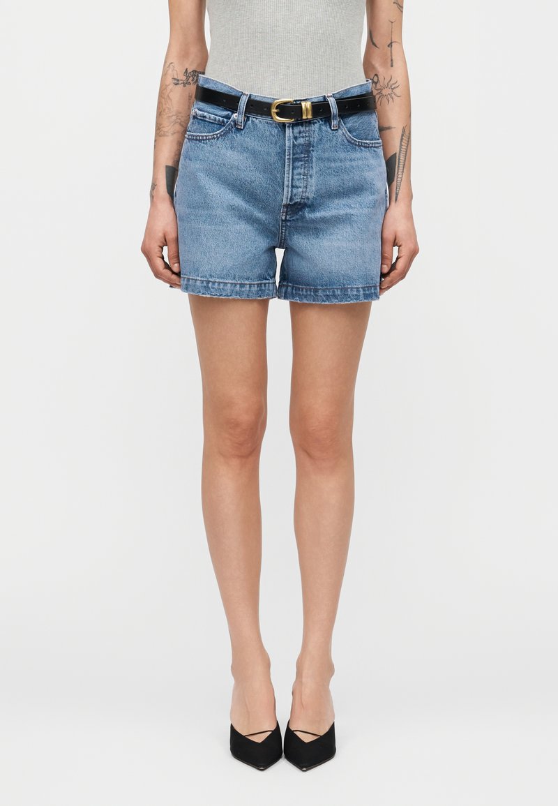 Γυναίκα που φοράει ψηλόμεσα μπλε τζιν σορτς με μαύρη ζώνη, λευκή αμάνικη μπλούζα, μαύρα μυτερά τακούνια και εμφανή τατουάζ στα μπράτσα.