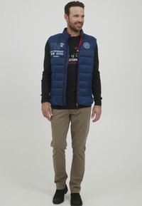 Temno modra puhvasta jopica s horizontalnim šivom, rdečimi detajli in logotipom. Nosi se čez črno srajco z dolgimi rokavi in kombinira z bež hlačami ter črnimi čevlji.