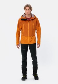 Oranje waterdichte jas met grijze accenten en een capuchon, gecombineerd met zwarte broek met getextureerde kniepaneel. Ontworpen voor buitensportactiviteiten.