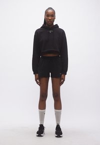 Γυναίκα στέκεται με το πρόσωπο προς τα εμπρός φορώντας μαύρο κοντό φούτερ με κουκούλα, μαύρο σορτς, λευκές κάλτσες μέχρι τον αστράγαλο και μαύρα αθλητικά παπούτσια σε απλό λευκό φόντο.