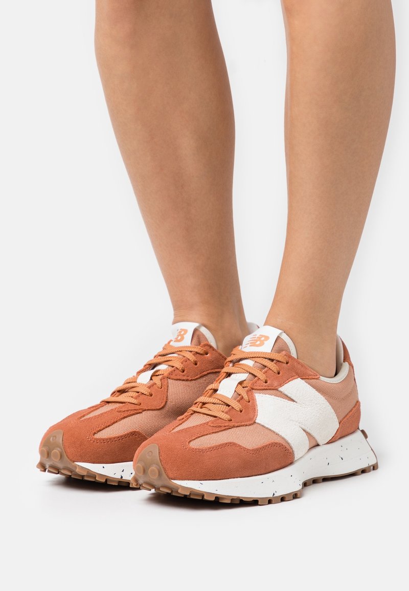Jambes portant des baskets New Balance orange et beige avec un logo "N" blanc et des semelles en gomme sur un fond blanc.