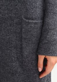 Szary płaszcz z filcu o teksturowanym wykończeniu, z przednią kieszenią i lekko luźnym krojem. Rękaw zakrywa część dłoni.