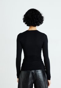 Persoon met kort, gekruld zwart haar, gekleed in een aansluitend zwart langemouwen shirt en zwarte leren broek, staand met de rug naar de camera op een witte achtergrond.