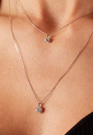 Colliers en argent avec de petits pendentifs ronds ornés de pierres claires. Un collier est plus haut que l'autre, mettant en avant un design superposé.