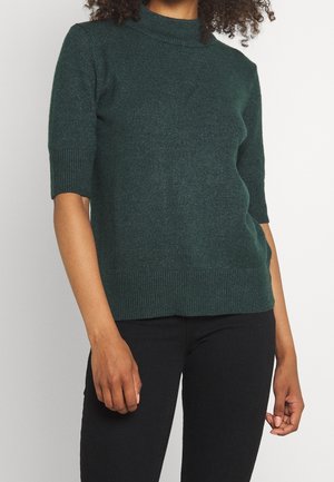 Femme portant un pull en maille vert foncé à manches courtes avec col et poignets côtelés, associé à un pantalon noir sur un fond uni.