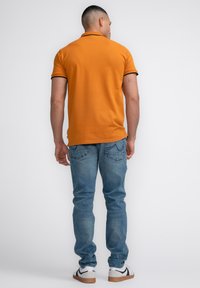 Oranžová polo tričko s černým lemem, spárované se světle modrými džíny. Model má na sobě bílé tenisky. Jednoduché pozadí.