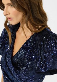 Marineblaues Paillettenkleid mit Wickeldesign, Puffärmeln und tiefem V-Ausschnitt; der Stoff hat eine strukturierte, funkelnde Oberfläche.