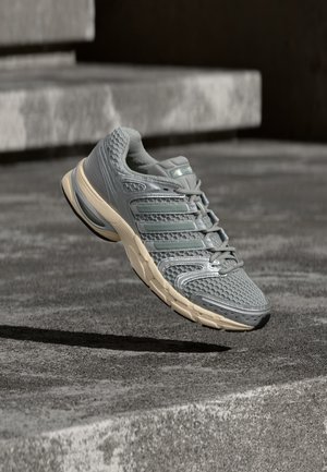 Šedá a béžová bežecká obuv Adidas, vo vzduchu nad sivými betónovými schodmi, s textúrou zo sieťoviny a odpruženou podrážkou.