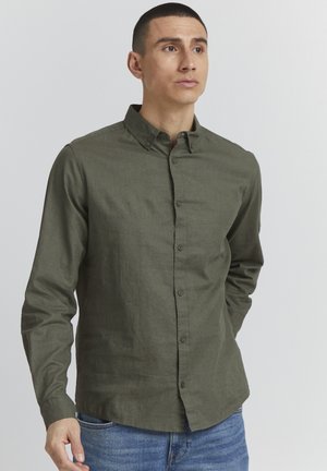 Long-sleeve button-up shirt in dark green textured fabric, featuring a pointed collar and front button closure. Worn with blue jeans.