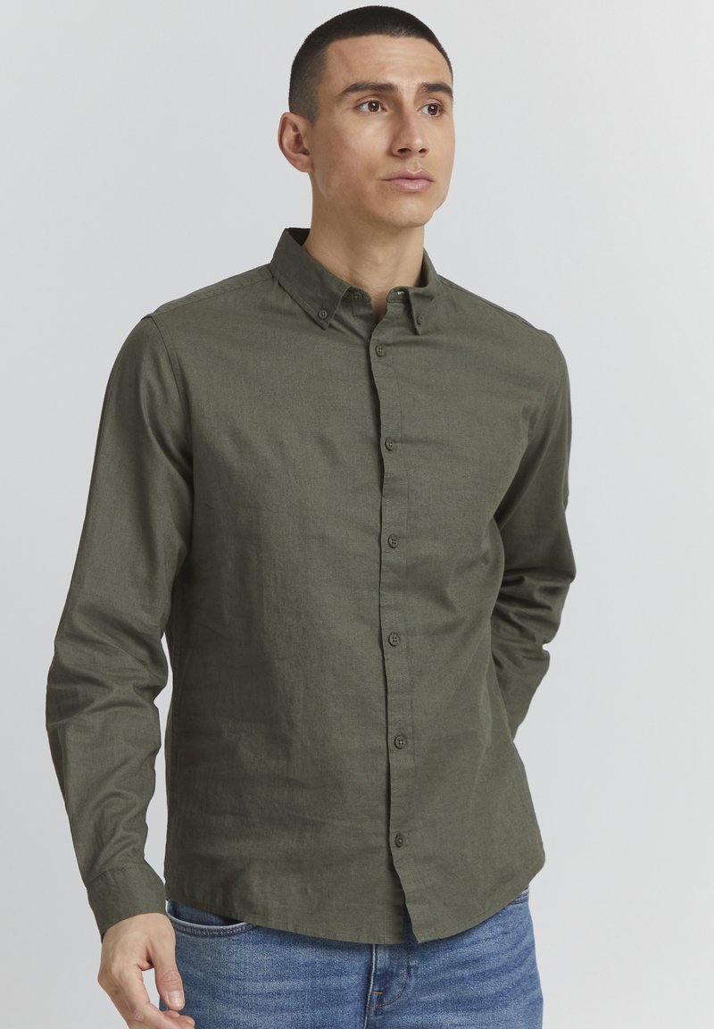 Langärmliges Button-up Hemd aus dunkelgrünem, strukturiertem Stoff, mit spitzem Kragen und vornem Knopfverschluss. Getragen mit blauen Jeans.