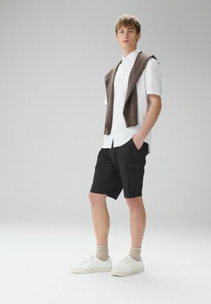 Młody mężczyzna stojący w białej koszuli z krótkim rękawem, czarnych spodenkach typu cargo, beżowych skarpetach, białych trampkach, z brązowym swetrem zarzuconym na ramiona.