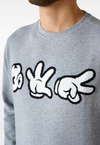 Sudadera gris con gráficos de manos bordados en blanco y negro texturizados, que incluyen un símbolo de paz y un gesto de pulgar hacia arriba, con un cuello acanalado.