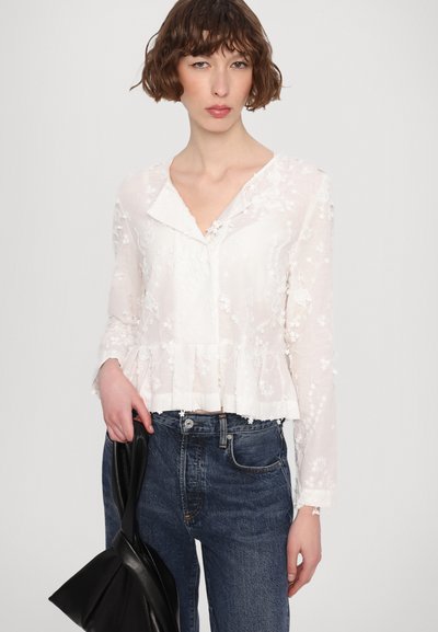 Bela vezenina bluza s peplum robom, z V-izrezom in dolgimi rokavi, v kombinaciji s temnimi kavbojkami in črno torbico.