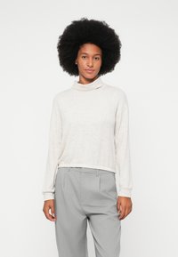 Woman with large afro hairstyle wearing a light beige turtleneck sweater and gray pinstriped pants, standing against a white background.