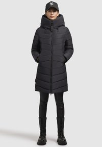 Manteau matelassé noir avec un col haut, fermeture éclair et deux poches latérales. Présente un matelassage horizontal et se marie avec des leggings noirs et des bottes.