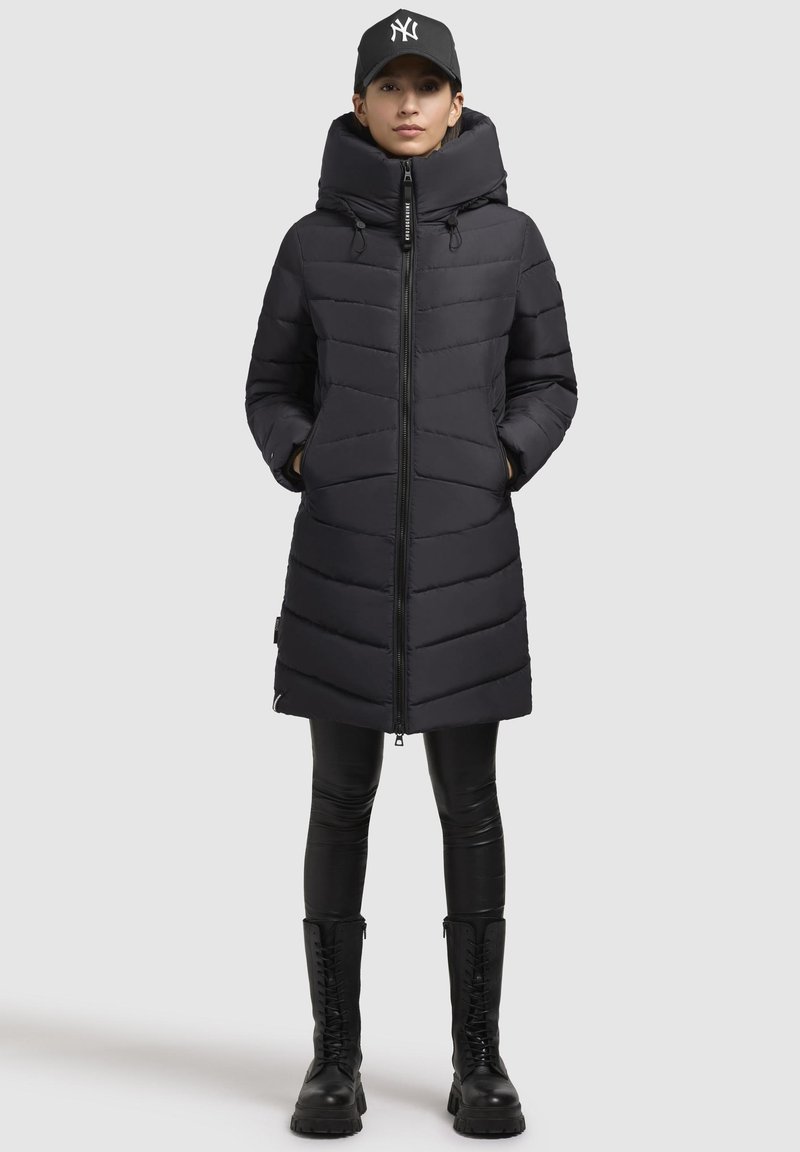 Manteau matelassé noir avec un col haut, fermeture éclair et deux poches latérales. Présente un matelassage horizontal et se marie avec des leggings noirs et des bottes.
