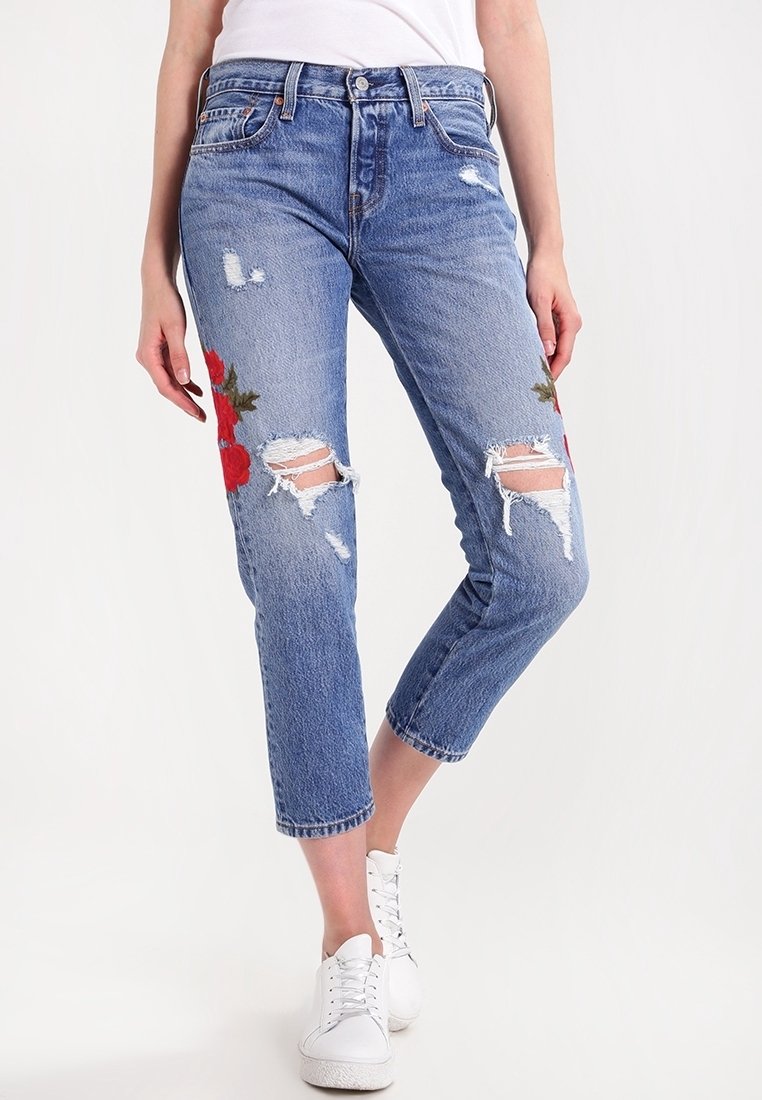 Jean court bleu avec genoux déchirés et broderie florale rouge, porté avec des baskets blanches et un haut blanc sur fond uni.