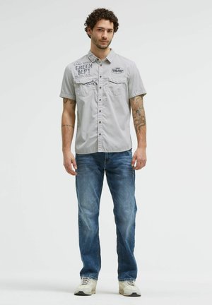 Junger Mann mit lockigem Haar steht und trägt ein hellgraues kurzärmeliges Hemd mit Knöpfen, blaue Jeans und weiße Sneakers vor einem einfarbigen Hintergrund.