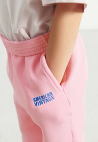 Ružové bavlnené tepláky s rebrovaným pásom, s bočnou vreckom a vyšitým modrým logom "AMERICAN VINTAGE" na nohe.