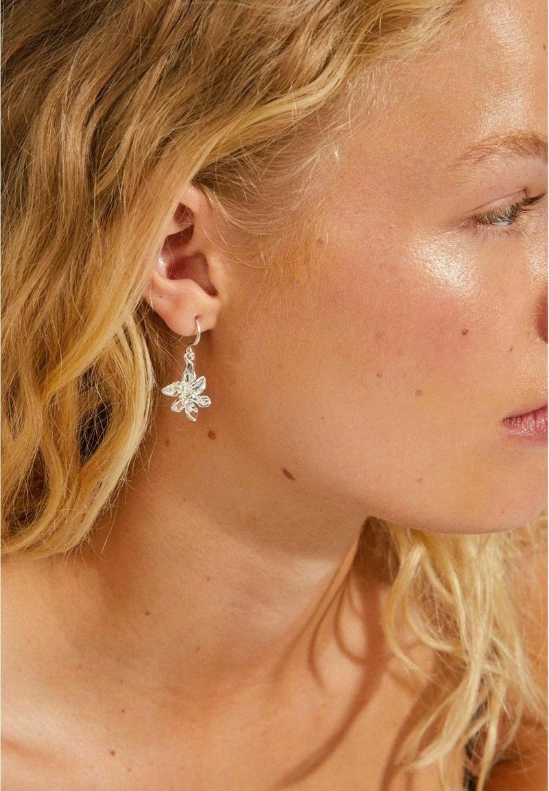 Silver floral earrings with a delicate design, featuring a blooming flower shape and a simple hoop, showcased against wavy blonde hair.