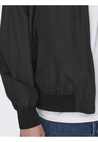 Black lightweight bomber jacket with elastic cuffs and hem, featuring a zip front and a smooth, plain texture. Worn over a white shirt.