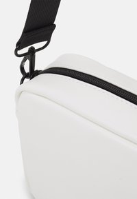 Borsa rettangolare bianca con superficie texturizzata, dotata di una cerniera nera e una tracolla nera regolabile. Vista ravvicinata che evidenzia i dettagli del design.