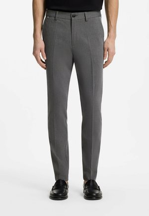 Homme portant un pantalon de costume gris ajusté avec pli avant, passants de ceinture noirs, chemise noire et mocassins en cuir noir sur fond blanc.