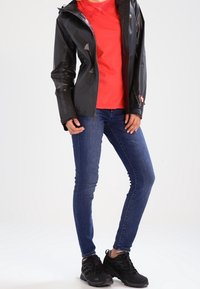 Sort vandtæt jakke, rød t-shirt, blå jeans og sorte sneakers. Jakken har hætte og lynlåse; jeansene er tætsiddende.