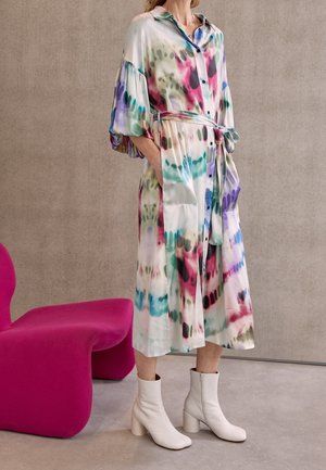 Femme portant une robe chemise midi tie-dye colorée avec des manches bouffantes et des bottines blanches, debout à côté d'une chaise moderne magenta.