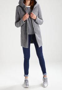 Persoon in een grijze hoodie-jack, gestreept shirt, donkere skinny jeans en zilveren sneakers, staand tegen een effen lichte achtergrond.