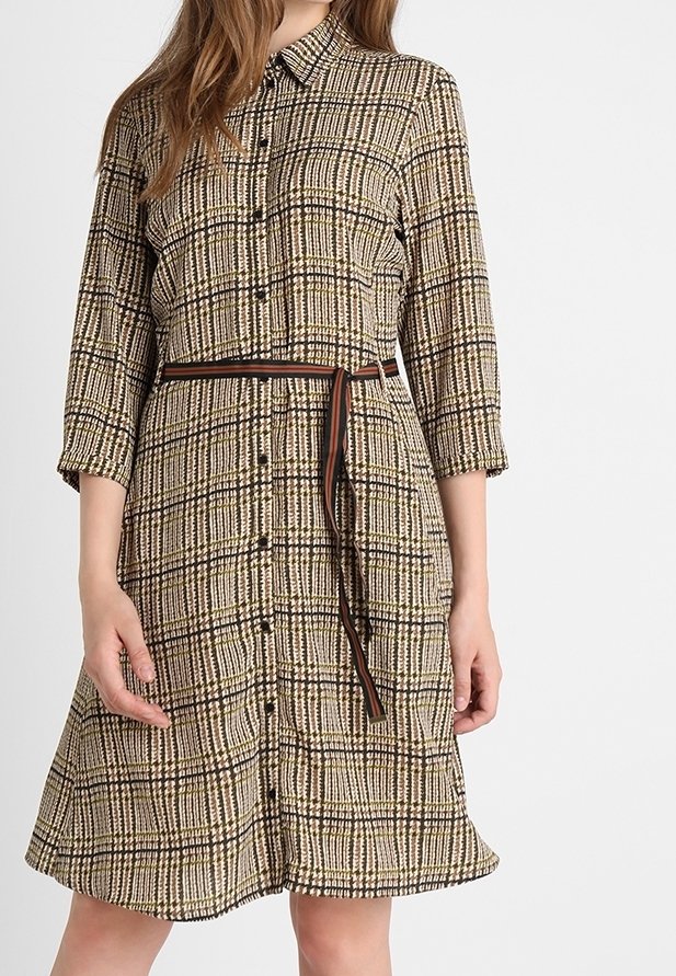Femme portant une robe à carreaux boutonnée, longueur genou, manches trois-quarts et fine ceinture, debout devant un fond uni.