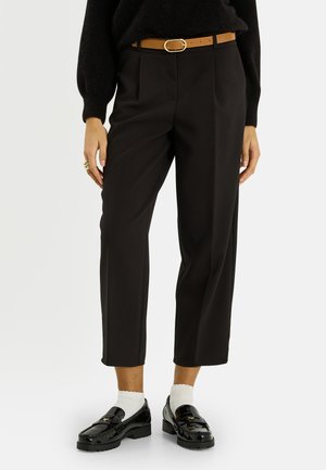 Femme portant un pantalon court noir, une ceinture beige, un pull noir, des chaussettes blanches et des mocassins en cuir verni noir sur un fond uni.