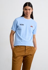 Svijetloplavi kratki rukav T-shirt s bijelim rebrastim ovratnikom, s crnim "ellesse" logotipom i malim crvenim i žutim dekorativnim detaljem.