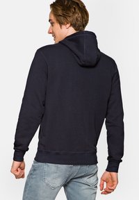 Granatowa bluza z kapturem ze sznurkiem, z ściągaczami na mankietach i u dołu. Posiada miękką fakturę oraz detale szycia na plecach.