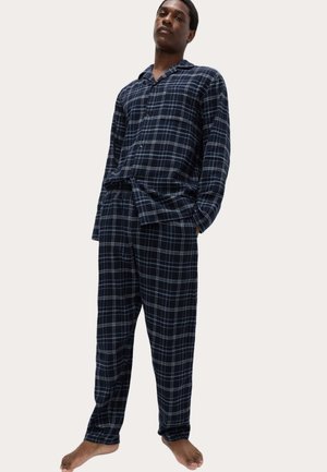 Mann steht barfuß und trägt ein marineblau-weiß kariertes langärmliges Pyjamashirt und passende Pyjamahosen vor einem neutralen Hintergrund.
