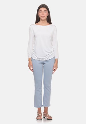 Donna in piedi con lunghi capelli scuri, che indossa una maglia bianca a maniche lunghe, pantaloni azzurri e sandali bianchi con tacco su uno sfondo semplice.