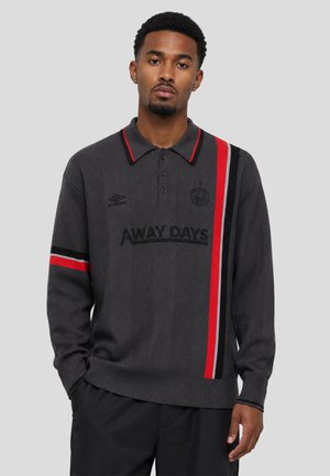 Uomo che indossa un maglione polo a maniche lunghe grigio scuro con strisce verticali rosse e nere e testo "AWAY DAYS" sul davanti su sfondo grigio.