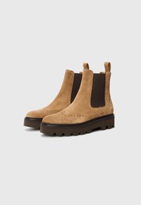 Bottines Chelsea en daim marron avec des panneaux élastiques latéraux, des détails en brogue et une semelle en caoutchouc noire épaisse. Boucles à l'arrière pour un enfilage facile.